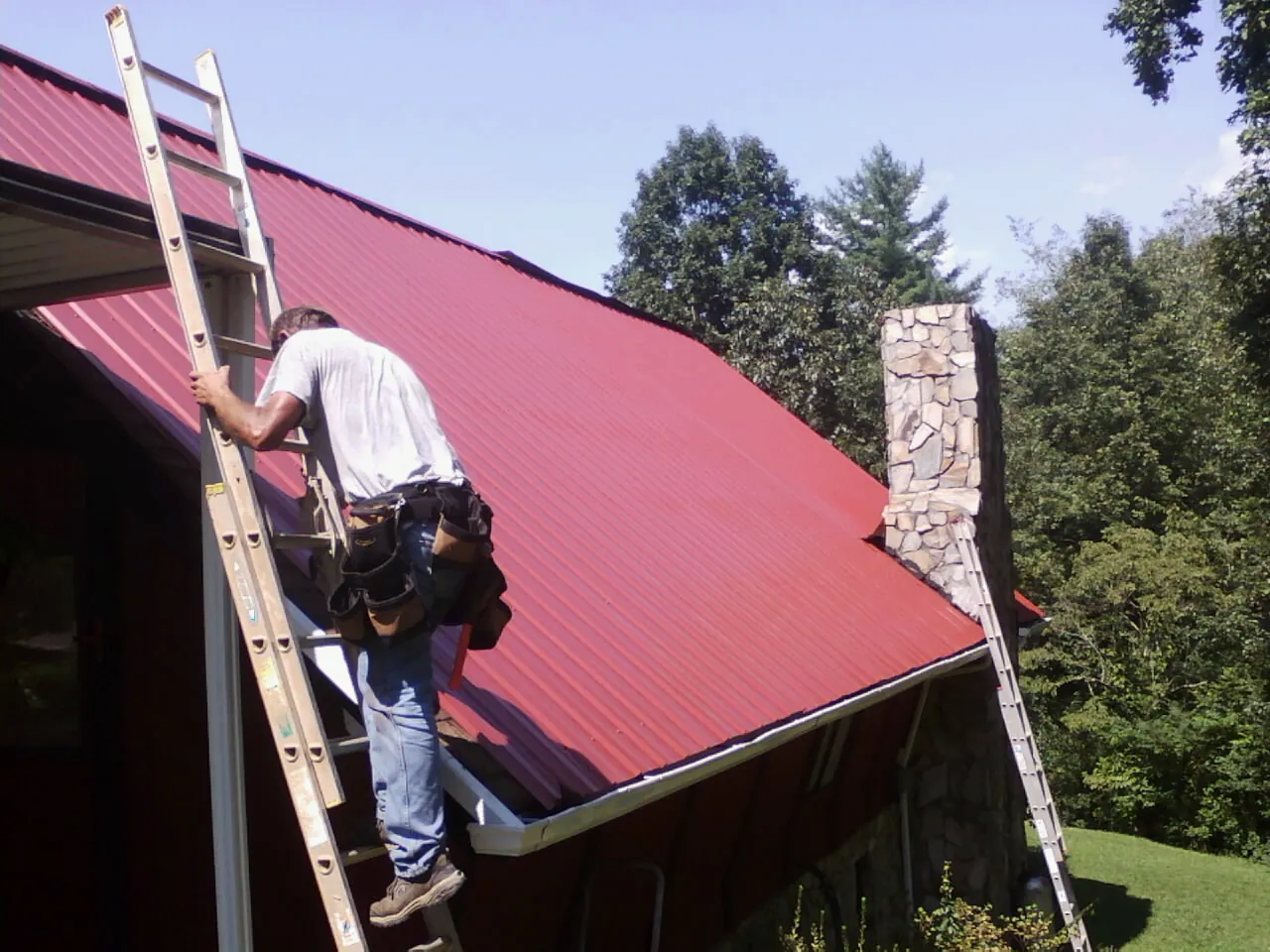 Roofer on a ladder beside a red metal roof and stone chimney — Soffit & Fascia Repair by Roofing Brothers in Centralia